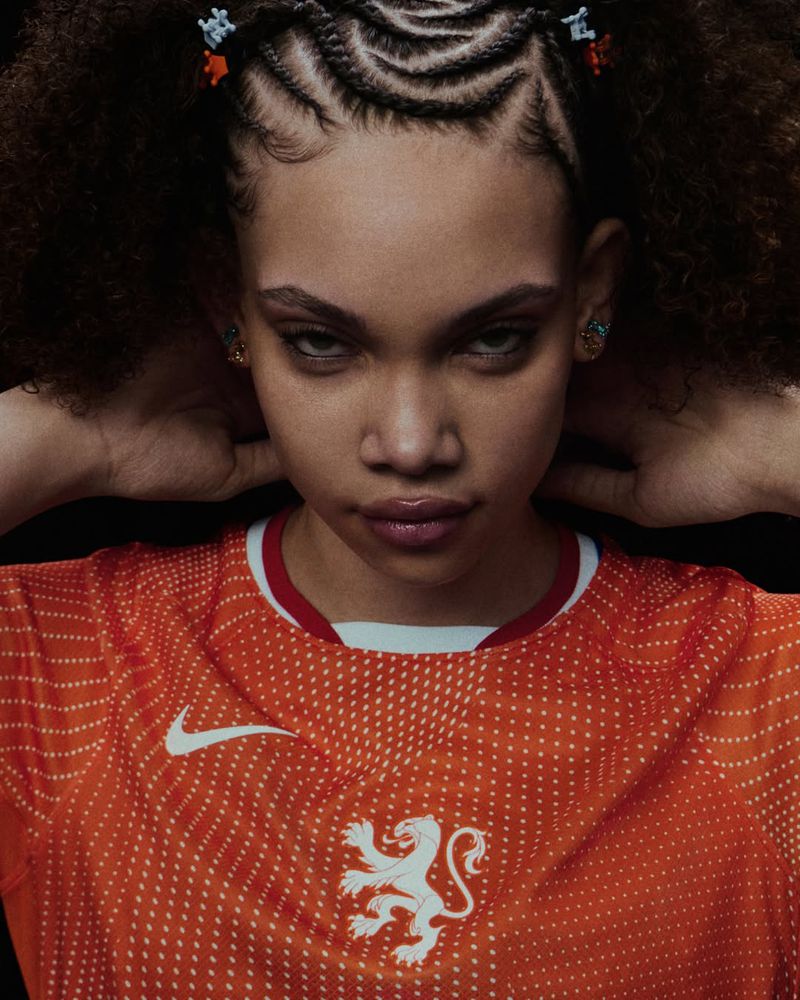 A young adult female with braided hair poses in a bright orange soccer jersey with a lion emblem against a dark background.