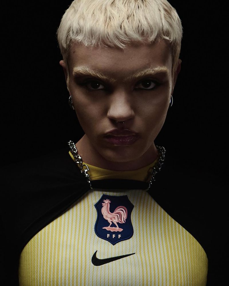 Close-up of a woman with blonde hair, wearing a yellow soccer jersey with the French national team emblem, against a black background.