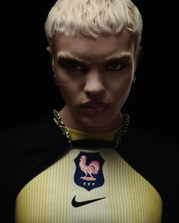 Close-up of a woman with blonde hair, wearing a yellow soccer jersey with the French national team emblem, against a black background.