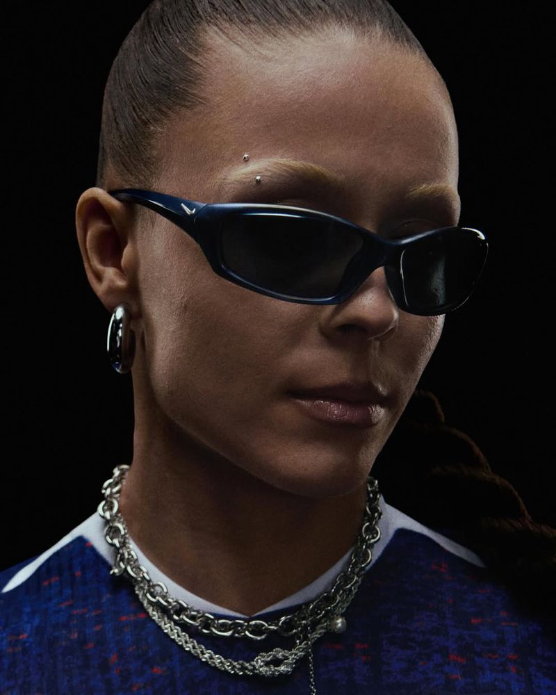 Woman with dark hair in a braid poses in a close-up studio shot wearing sunglasses, hoop earrings, and layered silver chains.