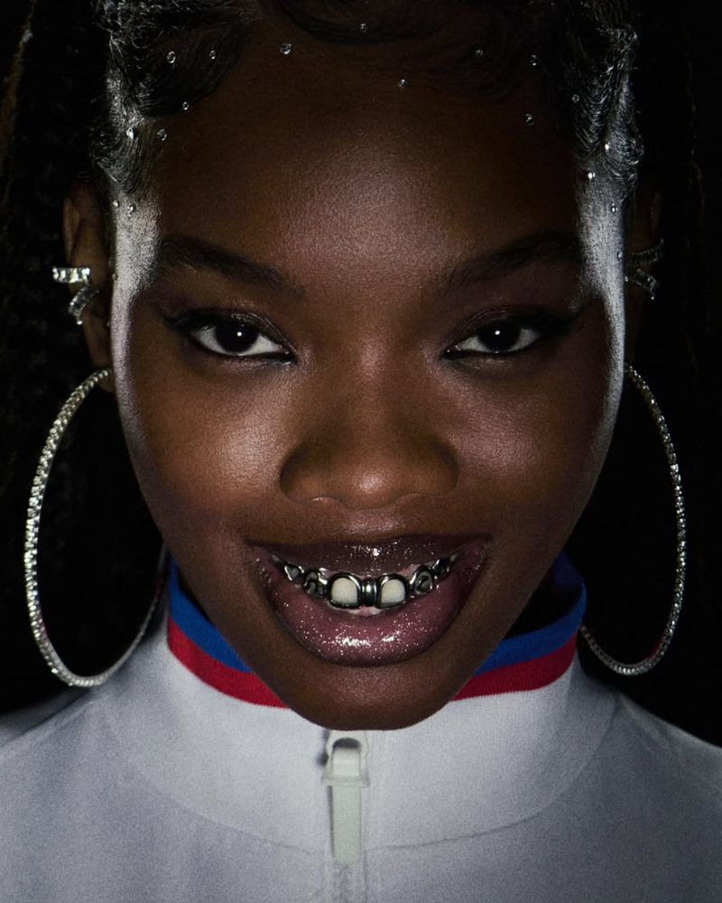 Close-up studio shot of an African-American woman smiling with dental grills, hoop earrings, and jeweled hair accents against a dark backdrop.