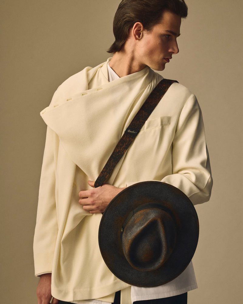 Portrait of a young adult male model with light skin wearing an off-white jacket, shoulder bag, and holding a dark-colored hat.