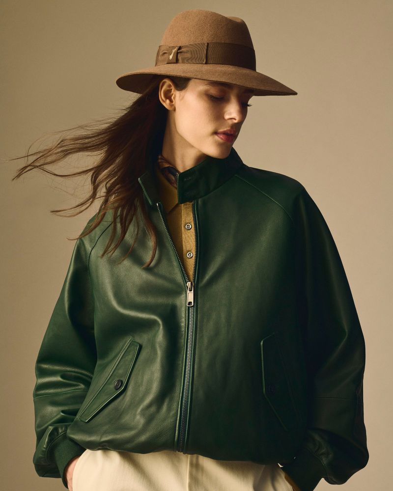 A woman in a green leather bomber jacket and brown hat poses in a studio with neutral lighting, showcasing fashion apparel.