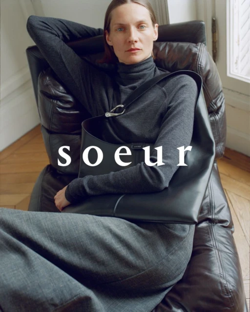 An adult female poses indoors, seated in a brown leather chair. She is wearing a gray turtleneck, carrying a black leather handbag. 