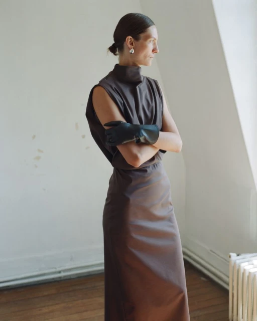 Elegant woman in a brown sleeveless dress and black gloves stands indoors in front of a window in a fashion-editorial shot.