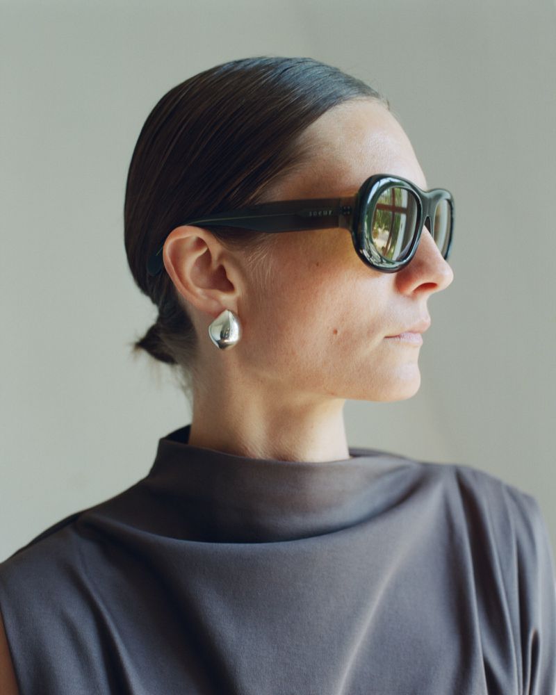 Adult woman with dark hair pulled back, wearing statement earrings and sunglasses, looking off-camera in a studio setting.