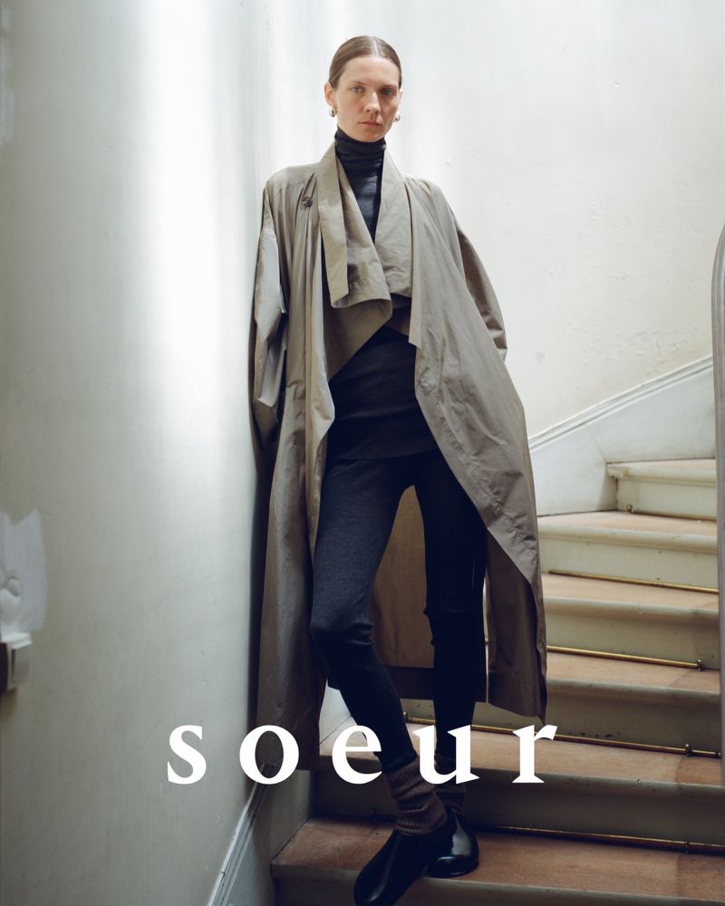 Fashion-forward woman poses on stairs in a flowing gray trench coat, black turtleneck, and leggings, captured in soft, natural light.