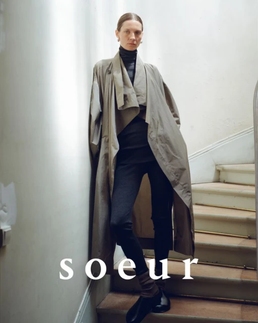Fashion-forward woman poses on stairs in a flowing gray trench coat, black turtleneck, and leggings, captured in soft, natural light.