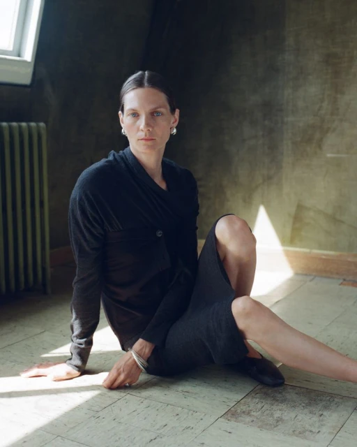 Elegant woman in a dark outfit sits on a marble floor near a radiator, looking directly at the camera with a neutral expression.