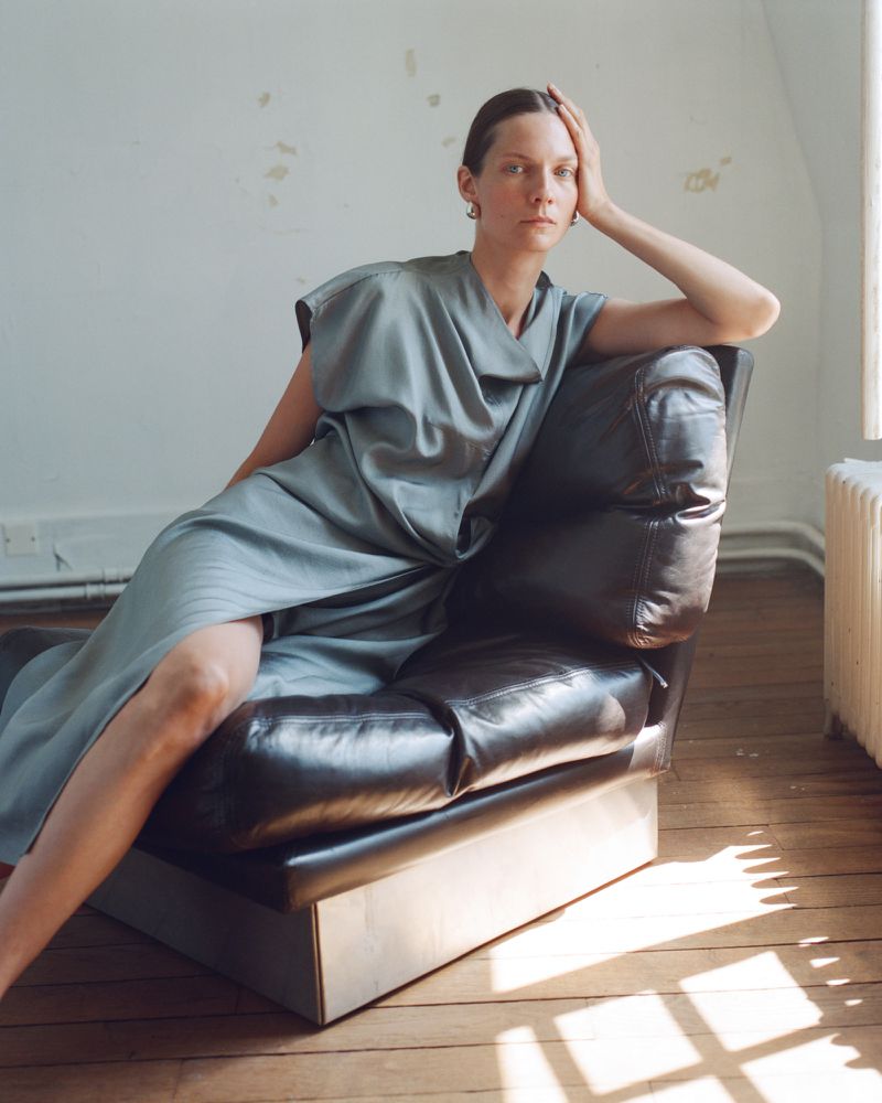 Adult woman in a blue dress sitting on a black leather chair indoors in front of a window with natural light, minimalist, and serene.
