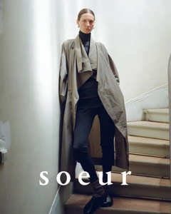 Model Karolin Wolter standing on an indoor staircase wearing an oversized beige trench coat, dark turtleneck, and black leggings.