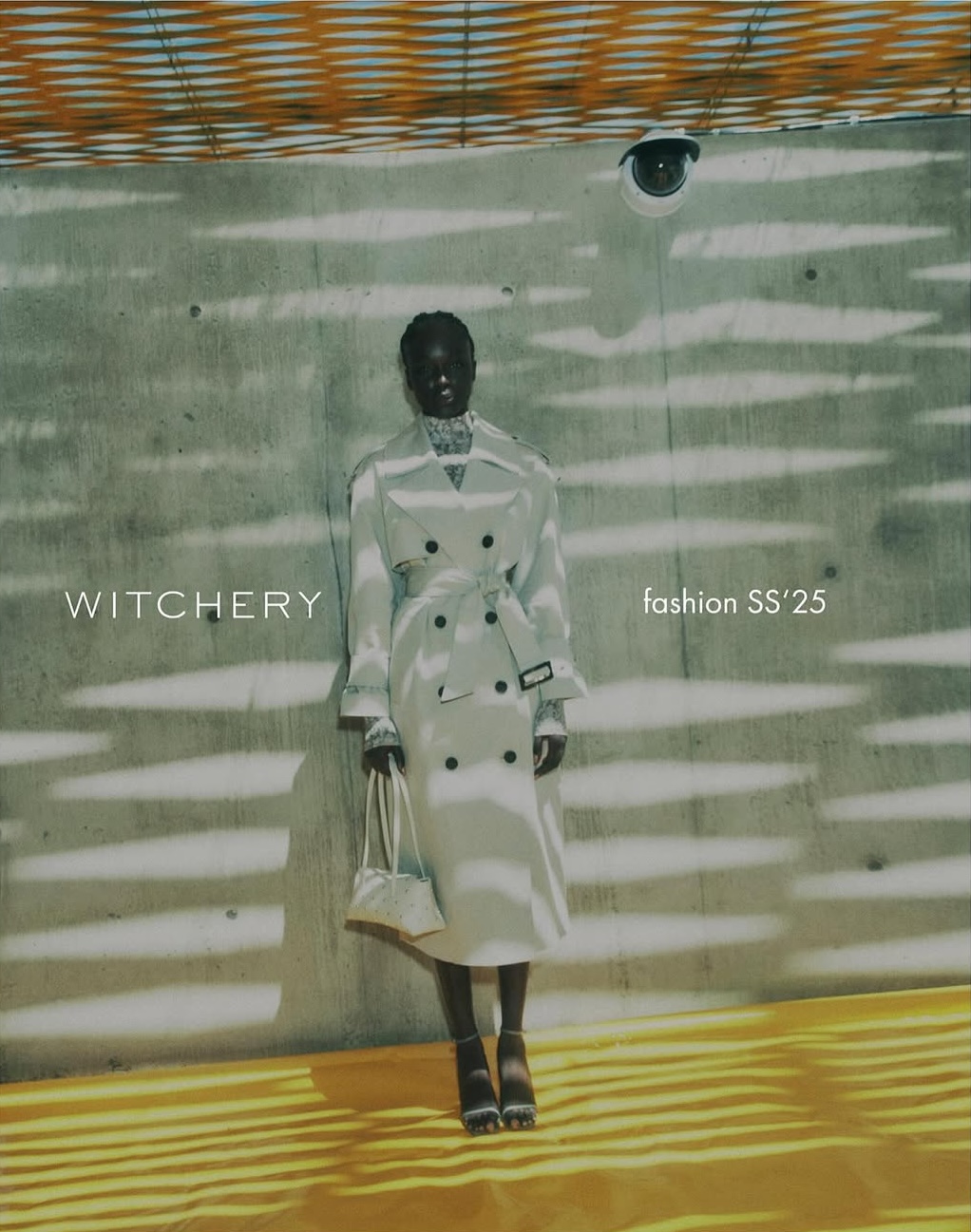 Fashion editorial featuring a Black woman in a white trench coat, holding a purse, posing against a textured concrete wall with shadows and light.