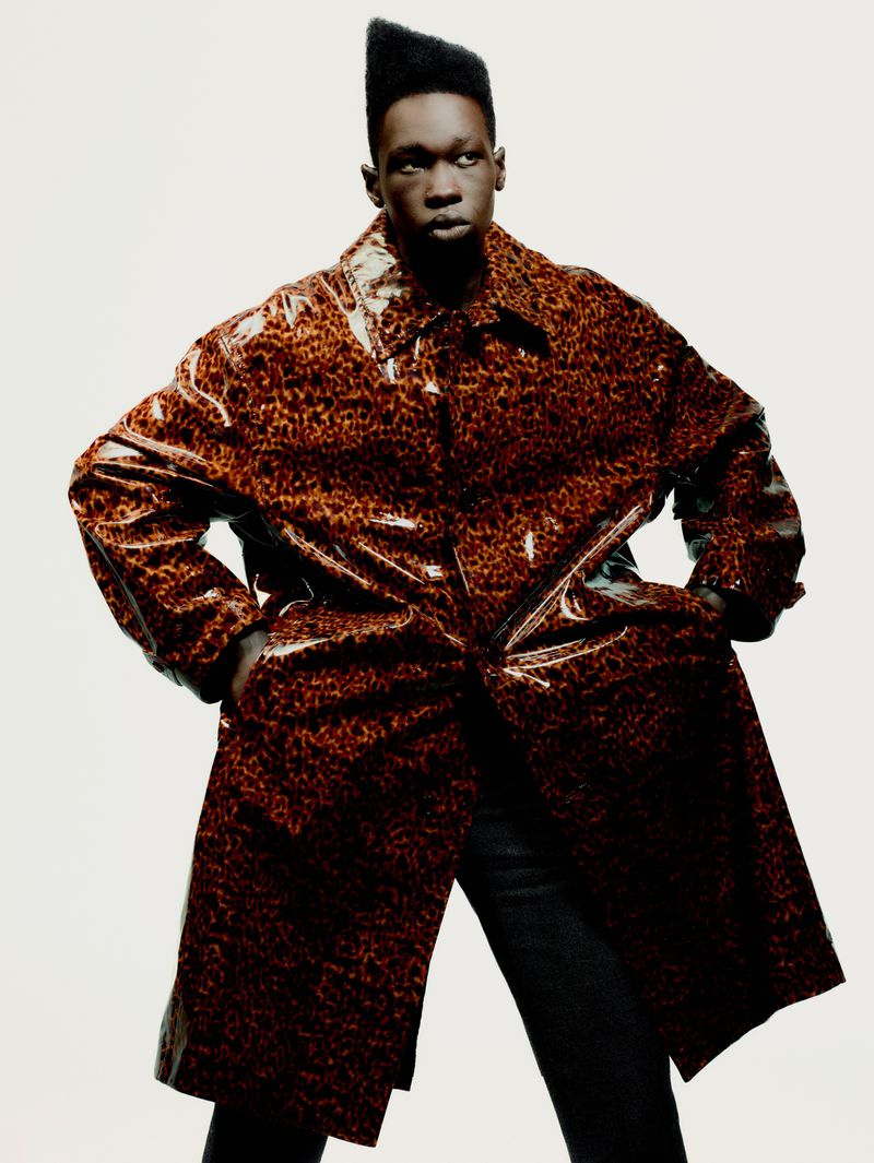 Adult male in a shiny brown and black leopard-print coat stands indoors against a plain white backdrop, hands in pockets, fashion portrait.