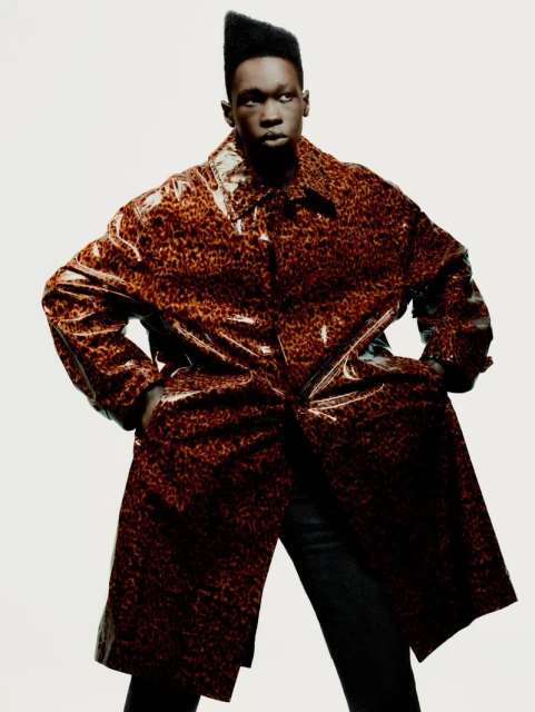 Adult male in a shiny brown and black leopard-print coat stands indoors against a plain white backdrop, hands in pockets, fashion portrait.