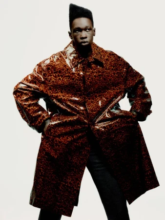 Adult male in a shiny brown and black leopard-print coat stands indoors against a plain white backdrop, hands in pockets, fashion portrait.