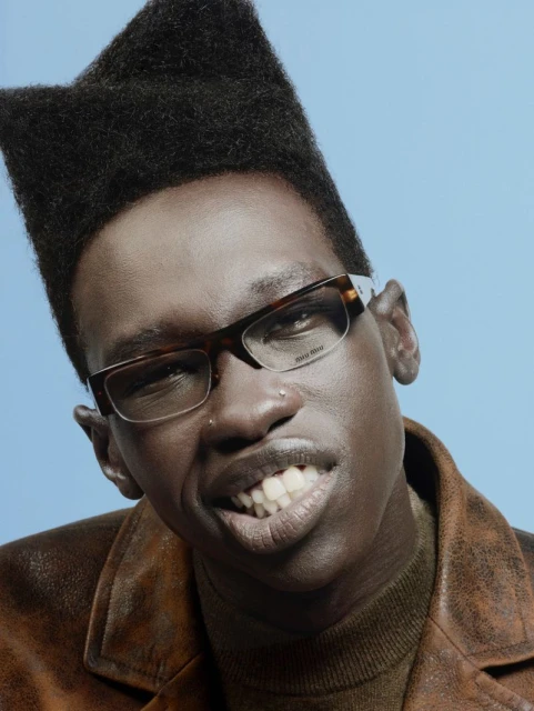 Portrait of a dark-skinned adult man with a distinctive high-top fade and tortoiseshell eyeglasses, smiling, against a light blue backdrop.