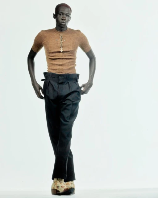 Stylish, dark-skinned man in a tan top and dark pants poses against a white backdrop, showcasing a high-fashion look.