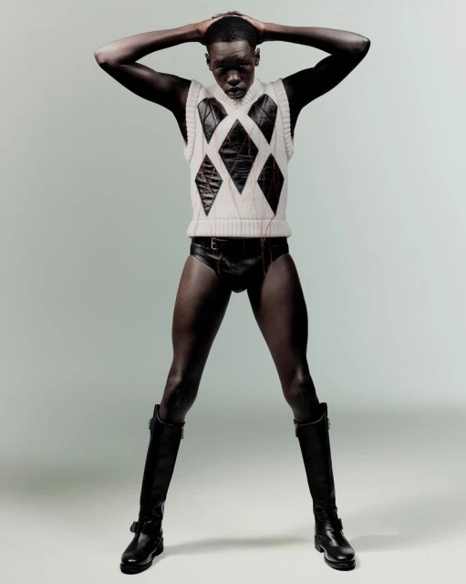 Studio shot of male fashion model with dark skin wearing a sleeveless argyle sweater, brief style bottoms, and knee-high boots in studio.