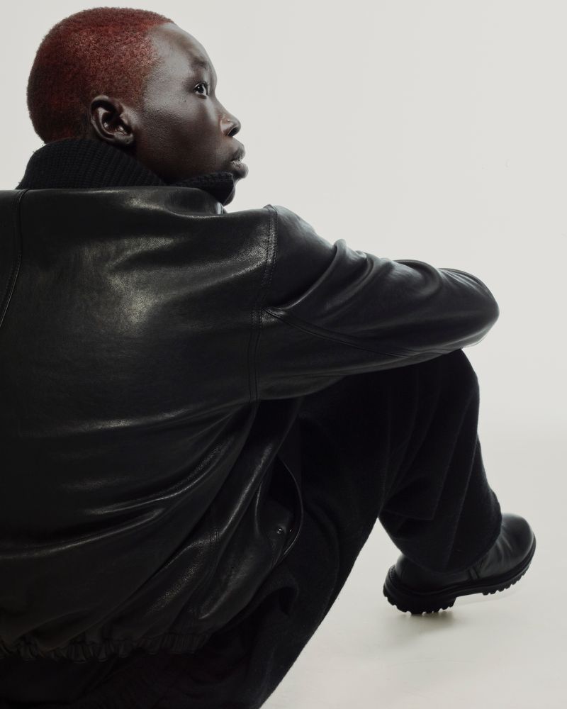 Portrait of a dark-skinned person with short red hair wearing a black leather jacket and sitting against a white backdrop.