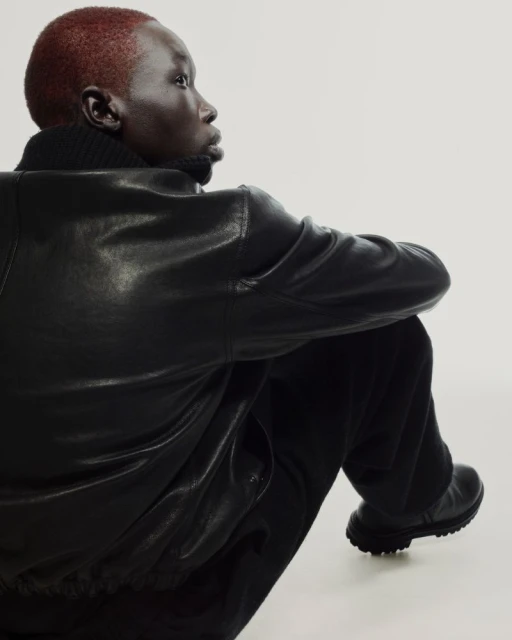 Portrait of a dark-skinned person with short red hair wearing a black leather jacket and sitting against a white backdrop.