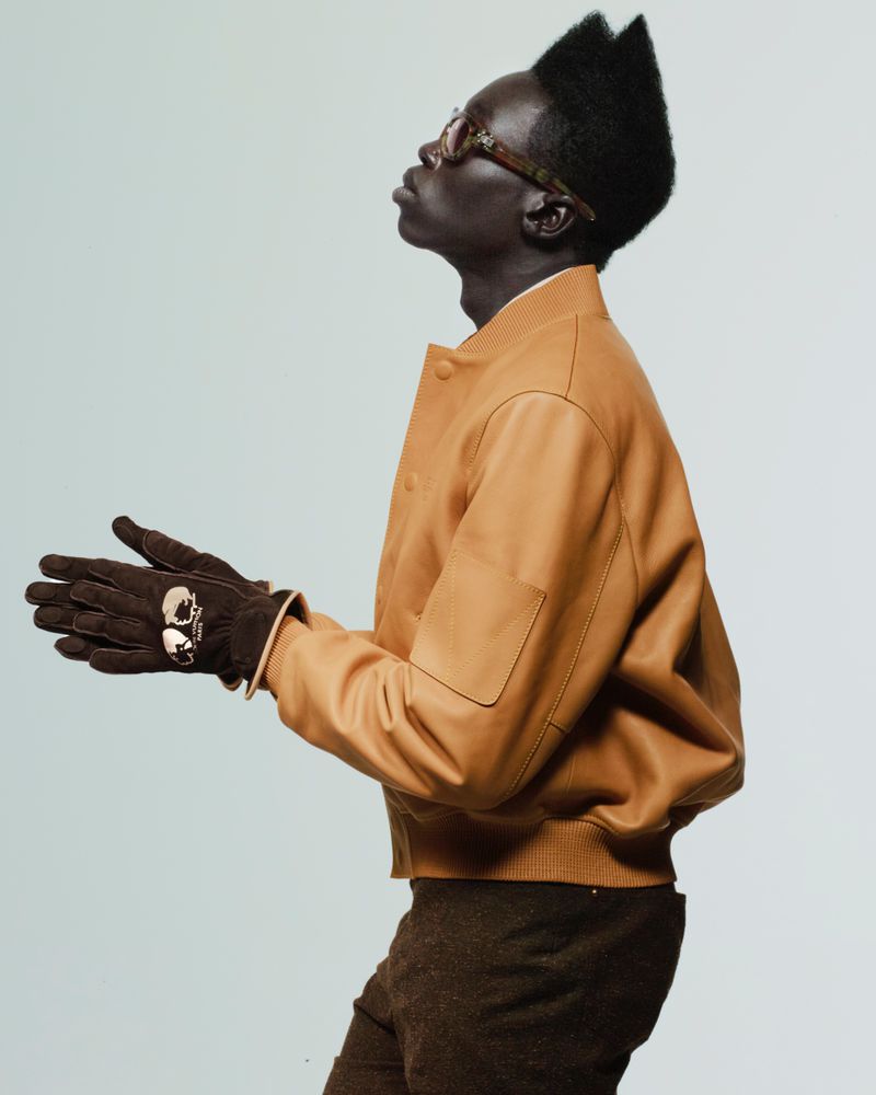 Side-view portrait of an adult male with a unique hairstyle wearing tan jacket, brown gloves, and glasses in front of a light background.