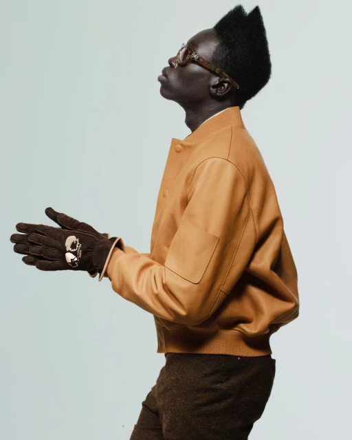 Side-view portrait of an adult male with a unique hairstyle wearing tan jacket, brown gloves, and glasses in front of a light background.