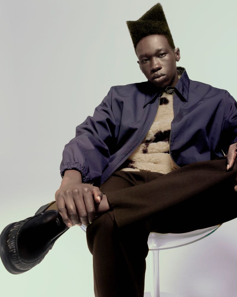 A Black male model wearing a dark blue jacket and brown pants is sitting on a clear chair in a studio with a neutral background.