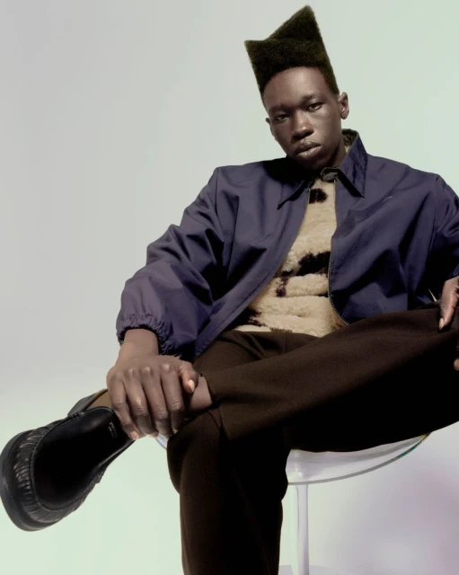 A Black male model wearing a dark blue jacket and brown pants is sitting on a clear chair in a studio with a neutral background.