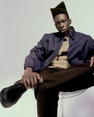 A Black male model wearing a dark blue jacket and brown pants is sitting on a clear chair in a studio with a neutral background.