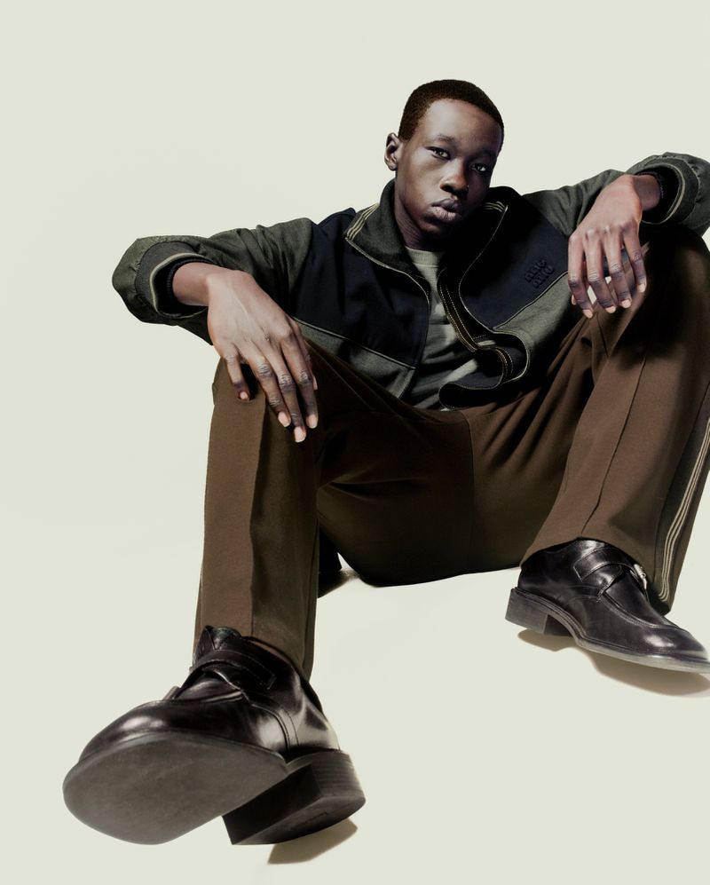 A young black man poses in a studio wearing a dark jacket, brown pants, and black shoes, with a neutral expression and a low-angle view.