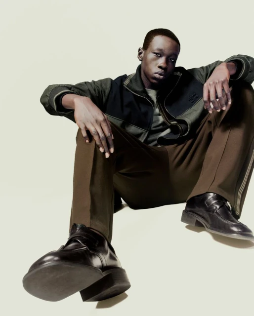 A young black man poses in a studio wearing a dark jacket, brown pants, and black shoes, with a neutral expression and a low-angle view.
