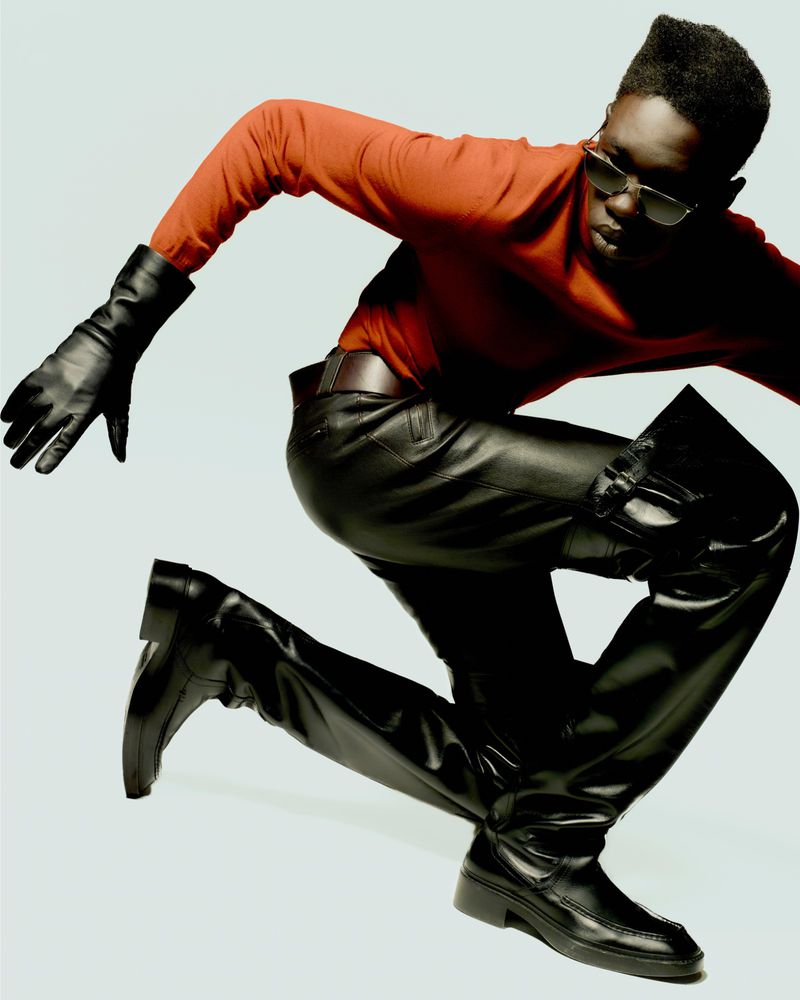 Fashion shot of a Black male model in an orange long-sleeved shirt, black leather pants, boots, and gloves, kneeling against a light background.
