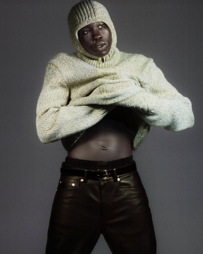 Fashionable dark-skinned model in a textured balaclava and sweater partially lifts the sweater to reveal their stomach in a studio setting.