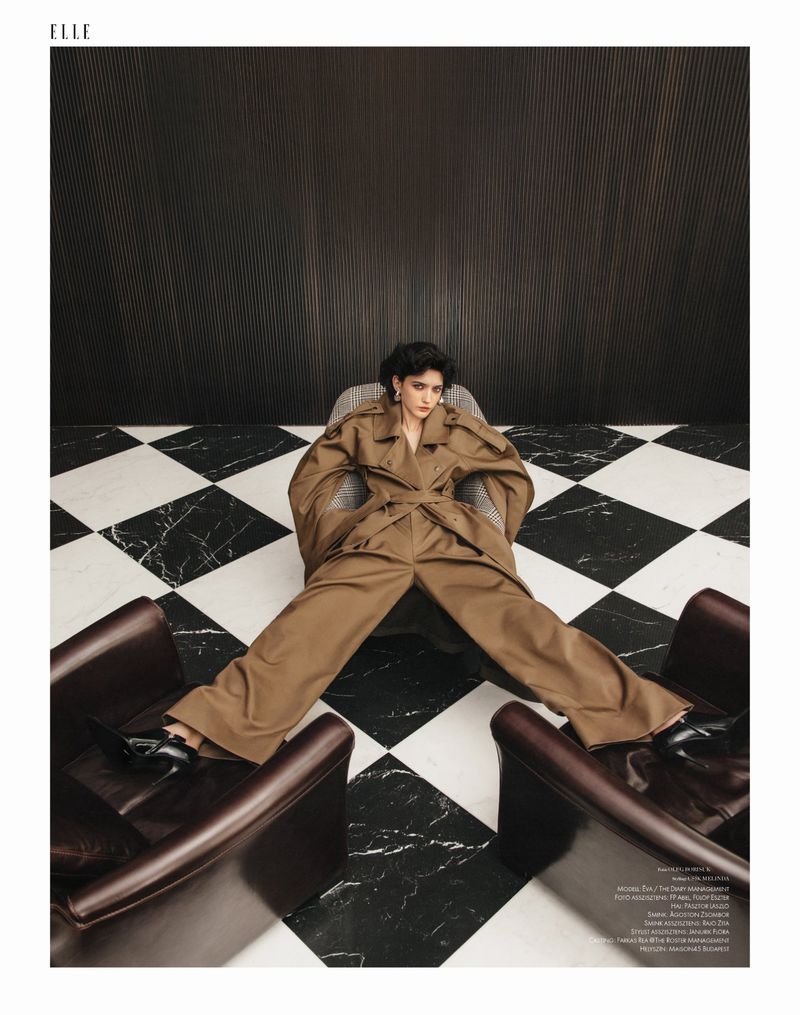 A young woman with short dark hair poses in a studio wearing a brown coat and pants, reclining between two brown leather armchairs.