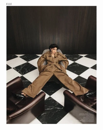 A young woman with short dark hair poses in a studio wearing a brown coat and pants, reclining between two brown leather armchairs.