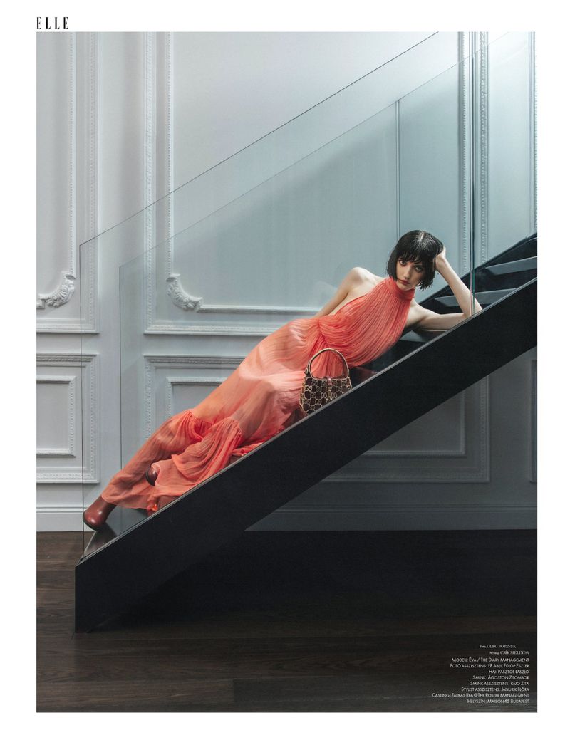A woman in a peach-colored dress is posed on a black staircase with a handbag. The setting is indoors with natural light.