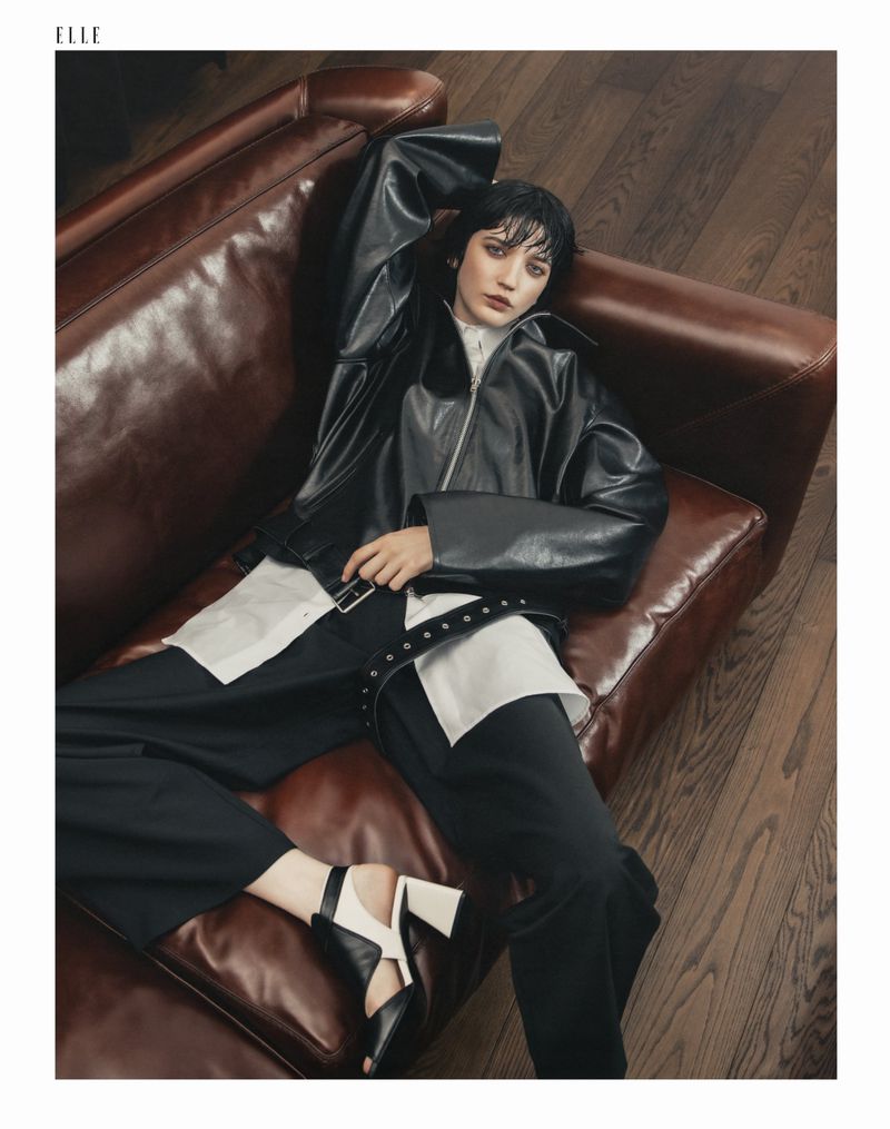 A woman lies on a brown leather couch wearing a black leather jacket, white shirt, and black pants, posing for a fashion editorial.