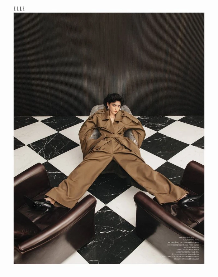 Model wearing a brown trench coat seated in a patterned armchair on a black and white tiled floor for Elle Hungary