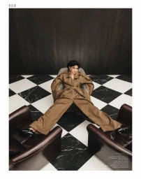 Model wearing a brown trench coat seated in a patterned armchair on a black and white tiled floor for Elle Hungary