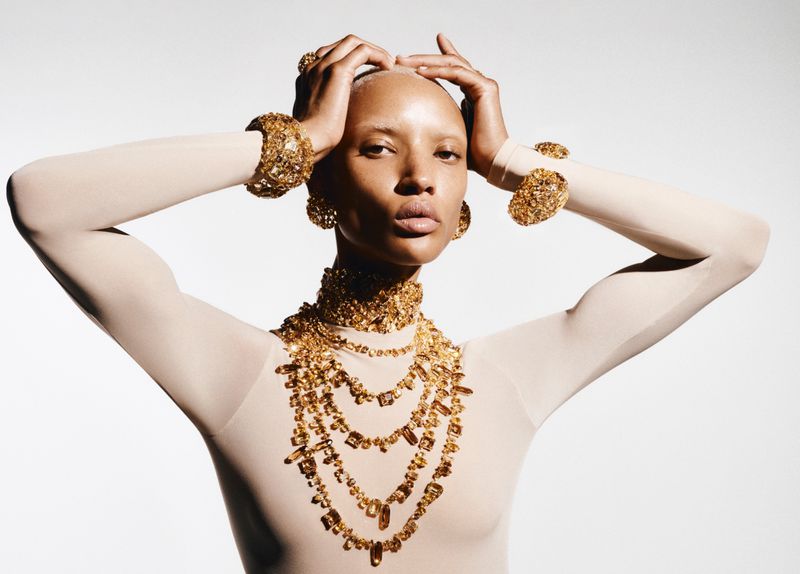 Woman adorned in gold jewelry, including a necklace, earrings, and bracelets, poses in a studio against a gradient white backdrop.