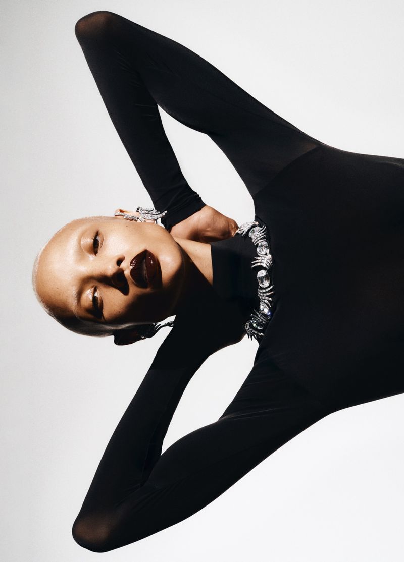 Fashion shot of a bald woman in a black bodysuit and striking jewellery, posing in front of a white background with dramatic lighting.