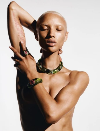 Close-up portrait of a bare-chested woman with short blonde hair, green and gold jeweled choker, bracelet, and ring on a white background.