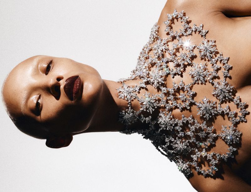 Glamour shot of a woman with bald head wearing a silver, snowflake-patterned decorative piece over her shoulders against a white background.
