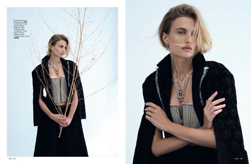 Fashion editorial featuring a woman with blonde hair in a corset top and black cape, posing with a dry branch against a light background.