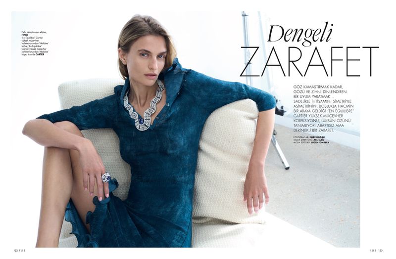 Elegant adult woman poses in a blue dress and statement jewelry while reclining on an off-white sofa in a bright, high-key indoor setting.