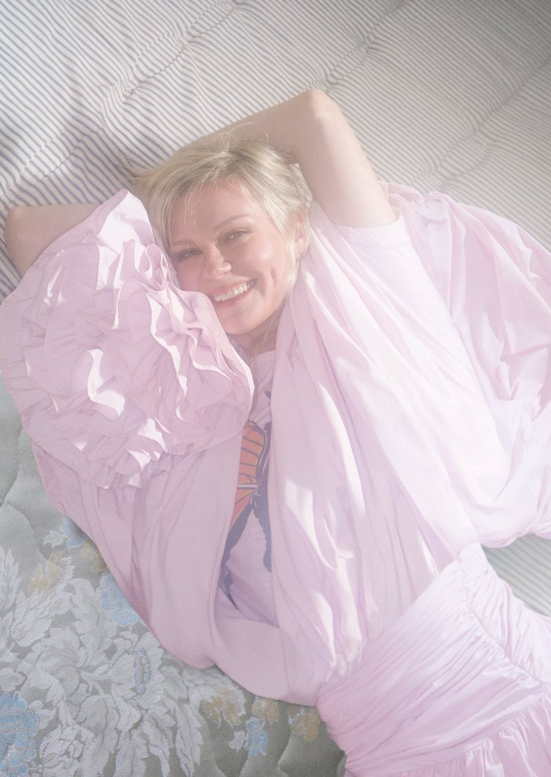 Smiling woman in a puffy lavender dress lying on a patterned mattress in a bright, soft-focus indoor setting.