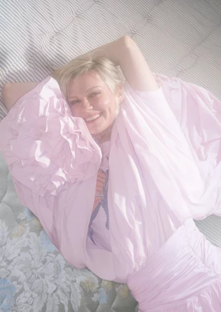 Smiling woman in a puffy lavender dress lying on a patterned mattress in a bright, soft-focus indoor setting.
