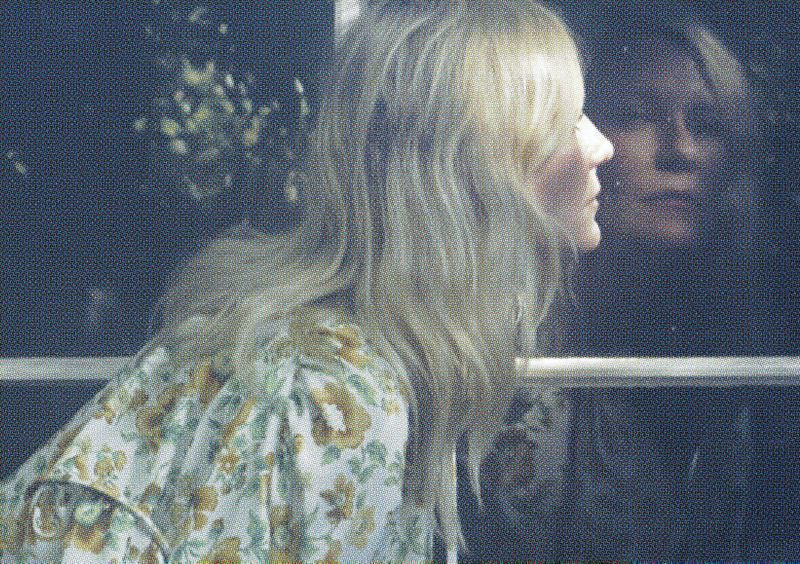 Blonde woman in floral top looks out window with her reflection visible. The scene is softly lit with muted colors and shallow focus.