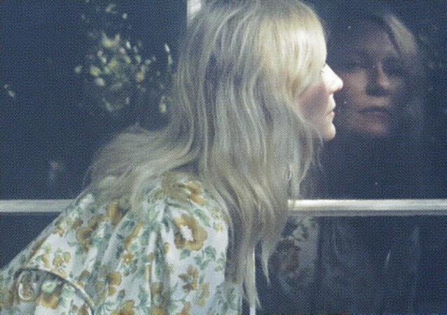 Blonde woman in floral top looks out window with her reflection visible. The scene is softly lit with muted colors and shallow focus.