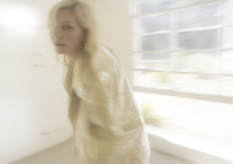 Blonde woman in a translucent garment stands in front of a window, soft focus, creating a dreamy, ethereal aesthetic.